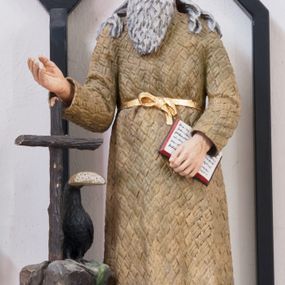 Zdjęcie nr 1: Rzeźba pełnoplastyczna umieszczona na niewielkim, prostopadłościennym postumencie, na którym ustawiona jest także skała zwieńczona drewnianym krzyżem oraz kruk z chlebem w dziobie. Święty Benedykt ukazany w pozycji stojącej, frontalnie. Został przedstawiony jako starszy mężczyzna o długich, siwych włosach oraz długiej, kędzierzawej brodzie w tym samym kolorze ubrany w długą, pustelniczą szatę przepasaną złotym sznurem. Głowa uniesiona ku górze, oczy szeroko otwarte. W lewej ręce święty trzyma otwartą księgę, prawą wyciąga przed siebie. Rzeźba w pełni polichromowana, pas złocony.