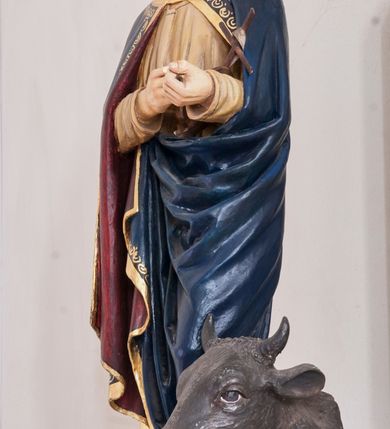 Zdjęcie nr 2: Rzeźba pełnoplastyczna, ustawiona na niewielkim, prostopadłościennym postumencie. Święty Łukasz Ewangelista ukazany  całej postaci, frontalnie, w pozycji stojącej. Został przedstawiony jako dojrzały mężczyzna o ciemnych, lekko falowanych włosach oraz gęstej, kędzierzawej brodzie w kolorze ciemnobrązowym. Twarz o regularnych rysach i wzroku uniesionym ku górze. Ręce ugięte w łokciu podtrzymujące niewielki, drewniany krzyż z banderolą. Ewangelista ubrany jest w beżową szatę o długich rękawach oraz obszerny ciemnogranatowy płaszcz z bordową podszewką i złoconą lamówką dekorowaną dodatkowo geometrycznym ornamentem. Spod szat widoczne są bose stopy postaci. U stóp symbol św. Łukasza Ewangelisty – wół leżącego na otwartej księdze z piórem. Figura w pełni polichromowana.