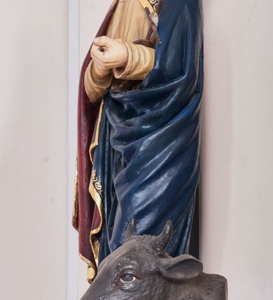 Zdjęcie nr 3: Rzeźba pełnoplastyczna, ustawiona na niewielkim, prostopadłościennym postumencie. Święty Łukasz Ewangelista ukazany  całej postaci, frontalnie, w pozycji stojącej. Został przedstawiony jako dojrzały mężczyzna o ciemnych, lekko falowanych włosach oraz gęstej, kędzierzawej brodzie w kolorze ciemnobrązowym. Twarz o regularnych rysach i wzroku uniesionym ku górze. Ręce ugięte w łokciu podtrzymujące niewielki, drewniany krzyż z banderolą. Ewangelista ubrany jest w beżową szatę o długich rękawach oraz obszerny ciemnogranatowy płaszcz z bordową podszewką i złoconą lamówką dekorowaną dodatkowo geometrycznym ornamentem. Spod szat widoczne są bose stopy postaci. U stóp symbol św. Łukasza Ewangelisty – wół leżącego na otwartej księdze z piórem. Figura w pełni polichromowana.