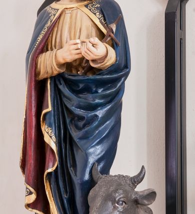 Zdjęcie nr 1: Rzeźba pełnoplastyczna, ustawiona na niewielkim, prostopadłościennym postumencie. Święty Łukasz Ewangelista ukazany  całej postaci, frontalnie, w pozycji stojącej. Został przedstawiony jako dojrzały mężczyzna o ciemnych, lekko falowanych włosach oraz gęstej, kędzierzawej brodzie w kolorze ciemnobrązowym. Twarz o regularnych rysach i wzroku uniesionym ku górze. Ręce ugięte w łokciu podtrzymujące niewielki, drewniany krzyż z banderolą. Ewangelista ubrany jest w beżową szatę o długich rękawach oraz obszerny ciemnogranatowy płaszcz z bordową podszewką i złoconą lamówką dekorowaną dodatkowo geometrycznym ornamentem. Spod szat widoczne są bose stopy postaci. U stóp symbol św. Łukasza Ewangelisty – wół leżącego na otwartej księdze z piórem. Figura w pełni polichromowana.
