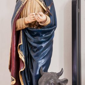 Zdjęcie nr 1: Rzeźba pełnoplastyczna, ustawiona na niewielkim, prostopadłościennym postumencie. Święty Łukasz Ewangelista ukazany  całej postaci, frontalnie, w pozycji stojącej. Został przedstawiony jako dojrzały mężczyzna o ciemnych, lekko falowanych włosach oraz gęstej, kędzierzawej brodzie w kolorze ciemnobrązowym. Twarz o regularnych rysach i wzroku uniesionym ku górze. Ręce ugięte w łokciu podtrzymujące niewielki, drewniany krzyż z banderolą. Ewangelista ubrany jest w beżową szatę o długich rękawach oraz obszerny ciemnogranatowy płaszcz z bordową podszewką i złoconą lamówką dekorowaną dodatkowo geometrycznym ornamentem. Spod szat widoczne są bose stopy postaci. U stóp symbol św. Łukasza Ewangelisty – wół leżącego na otwartej księdze z piórem. Figura w pełni polichromowana.