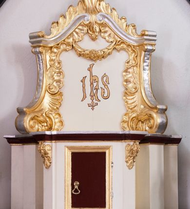 Zdjęcie nr 2: Tabernakulum z tronem wystawienia uskokowe, ustawione na profilowanym cokole, ujęte ustawionymi ukośnie zwielokrotnionymi filarami, zamknięte wydatnym, profilowanym gzymsem wyłamanym w osiach podpór. Profile gzymsu złocone, srebrzone oraz malowane na kolor ciemnobrązowy. Pierwsza para filarów w górnej części dekorowana złoconymi wolutami z motywem palmet oraz rozet. Drzwiczki prostokątne, ujęte profilowaną, złoconą ramą z płyciną malowaną w kolorze ciemnobrązowym. Zwieńczenie ujęte dwoma ustawionymi ukośnie spływami wolutowymi dekorowanymi rocaille&#039;ami, dźwigające przerwany przyczółek o falistej formie z dekoracją rocaille&#039;ów oraz girland. W polu zwieńczenia malowany monogram IHS. Struktura tabernakulum malowana w kolorze jasnobeżowym, ornamentyka złocona i srebrzona. Detale z kolorze ciemnego brązu i szarości.