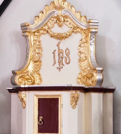 Zdjęcie nr 1: Tabernakulum z tronem wystawienia uskokowe, ustawione na profilowanym cokole, ujęte ustawionymi ukośnie zwielokrotnionymi filarami, zamknięte wydatnym, profilowanym gzymsem wyłamanym w osiach podpór. Profile gzymsu złocone, srebrzone oraz malowane na kolor ciemnobrązowy. Pierwsza para filarów w górnej części dekorowana złoconymi wolutami z motywem palmet oraz rozet. Drzwiczki prostokątne, ujęte profilowaną, złoconą ramą z płyciną malowaną w kolorze ciemnobrązowym. Zwieńczenie ujęte dwoma ustawionymi ukośnie spływami wolutowymi dekorowanymi rocaille'ami, dźwigające przerwany przyczółek o falistej formie z dekoracją rocaille'ów oraz girland. W polu zwieńczenia malowany monogram IHS. Struktura tabernakulum malowana w kolorze jasnobeżowym, ornamentyka złocona i srebrzona. Detale z kolorze ciemnego brązu i szarości.