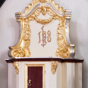 Zdjęcie nr 1: Tabernakulum z tronem wystawienia uskokowe, ustawione na profilowanym cokole, ujęte ustawionymi ukośnie zwielokrotnionymi filarami, zamknięte wydatnym, profilowanym gzymsem wyłamanym w osiach podpór. Profile gzymsu złocone, srebrzone oraz malowane na kolor ciemnobrązowy. Pierwsza para filarów w górnej części dekorowana złoconymi wolutami z motywem palmet oraz rozet. Drzwiczki prostokątne, ujęte profilowaną, złoconą ramą z płyciną malowaną w kolorze ciemnobrązowym. Zwieńczenie ujęte dwoma ustawionymi ukośnie spływami wolutowymi dekorowanymi rocaille'ami, dźwigające przerwany przyczółek o falistej formie z dekoracją rocaille'ów oraz girland. W polu zwieńczenia malowany monogram IHS. Struktura tabernakulum malowana w kolorze jasnobeżowym, ornamentyka złocona i srebrzona. Detale z kolorze ciemnego brązu i szarości.
