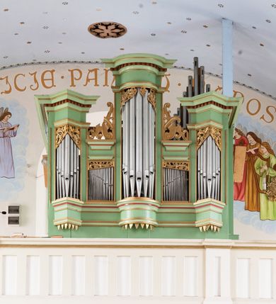 Zdjęcie nr 1: Prospekt organowy jednokondygnacyjny, pięcioosiowy o formach architektonicznych umieszczony na wysokim cokole.
Osie nieparzyste (pierwsza, trzecia i piąta) z większymi piszczałkami wypukłe, a pierwsza i piąta, na rzucie trójkąta niższe od środkowej. Wszystkie zwieńczone pełnym belkowaniem z wydatnym, profilowanym gzymsem dekorowanym kostkowaniem. W górnej części złocony, ażurowy lambrekin utworzony z ornamentu małżowinowo-chrząstkowego. Osie parzyste są niższe z płaskimi, prostokątnymi niszami mieszczącymi mniejsze piszczałki. Wieńczą je profilowano-fazowane konsole oraz ażurowe struktury z ornamentu małżowinowo-chrząstkowego. Prospekt polichromowany w odcieniach seledynowej zieleni z czerwonymi detalami, zaś ornamentyka i załamania profili architektonicznych złocone.