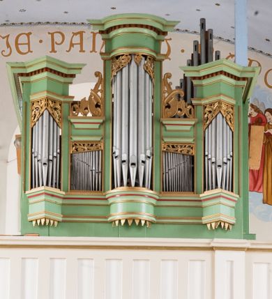 Zdjęcie nr 2: Prospekt organowy jednokondygnacyjny, pięcioosiowy o formach architektonicznych umieszczony na wysokim cokole.
Osie nieparzyste (pierwsza, trzecia i piąta) z większymi piszczałkami wypukłe, a pierwsza i piąta, na rzucie trójkąta niższe od środkowej. Wszystkie zwieńczone pełnym belkowaniem z wydatnym, profilowanym gzymsem dekorowanym kostkowaniem. W górnej części złocony, ażurowy lambrekin utworzony z ornamentu małżowinowo-chrząstkowego. Osie parzyste są niższe z płaskimi, prostokątnymi niszami mieszczącymi mniejsze piszczałki. Wieńczą je profilowano-fazowane konsole oraz ażurowe struktury z ornamentu małżowinowo-chrząstkowego. Prospekt polichromowany w odcieniach seledynowej zieleni z czerwonymi detalami, zaś ornamentyka i załamania profili architektonicznych złocone.