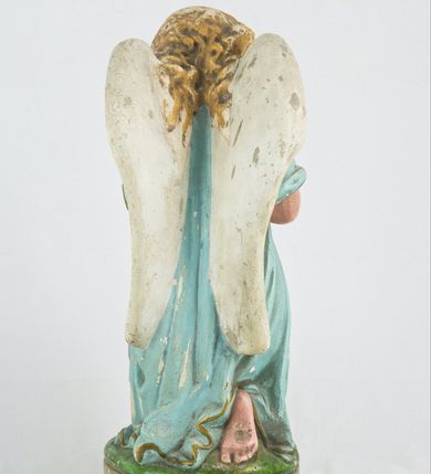 Zdjęcie nr 4: Rzeźba przedstawia anioła klęczącego na lewym kolanie na cokole imitującym rosnącą trawę. Postać ma ręce złożone do modlitwy i opuszczoną głowę z szeroką twarzą o jasnych oczach, małym nosie i czerwonych, niewielkich ustach; jasne włosy opadają jej na plecy. Jest ubrana w niebieską suknię ze złotą lamówką i kołnierzykiem w kropki; skrzydła białe, złożone. 