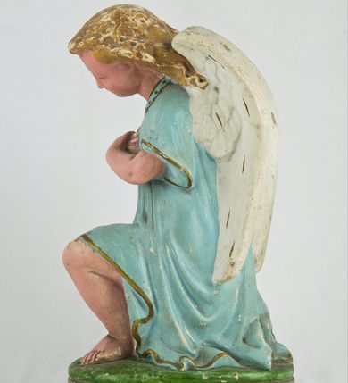 Zdjęcie nr 3: Rzeźba przedstawia anioła klęczącego na lewym kolanie na cokole imitującym rosnącą trawę. Postać ma ręce złożone do modlitwy i opuszczoną głowę z szeroką twarzą o jasnych oczach, małym nosie i czerwonych, niewielkich ustach; jasne włosy opadają jej na plecy. Jest ubrana w niebieską suknię ze złotą lamówką i kołnierzykiem w kropki; skrzydła białe, złożone. 