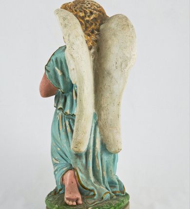 Zdjęcie nr 5: Rzeźba przedstawia anioła klęczącego na lewym kolanie na cokole imitującym rosnącą trawę. Postać ma ręce złożone do modlitwy i opuszczoną głowę z szeroką twarzą o jasnych oczach, małym nosie i czerwonych, niewielkich ustach; jasne włosy opadają jej na plecy. Jest ubrana w niebieską suknię ze złotą lamówką i kołnierzykiem w kropki; skrzydła białe, złożone. 