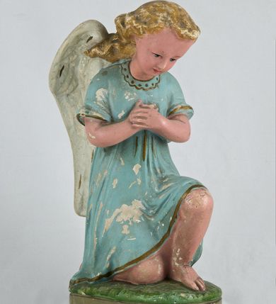 Zdjęcie nr 2: Rzeźba przedstawia anioła klęczącego na lewym kolanie na cokole imitującym rosnącą trawę. Postać ma ręce złożone do modlitwy i opuszczoną głowę z szeroką twarzą o jasnych oczach, małym nosie i czerwonych, niewielkich ustach; jasne włosy opadają jej na plecy. Jest ubrana w niebieską suknię ze złotą lamówką i kołnierzykiem w kropki; skrzydła białe, złożone. 