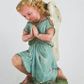 Zdjęcie nr 1: Rzeźba przedstawia anioła klęczącego na lewym kolanie na cokole imitującym rosnącą trawę. Postać ma ręce złożone do modlitwy i opuszczoną głowę z szeroką twarzą o jasnych oczach, małym nosie i czerwonych, niewielkich ustach; jasne włosy opadają jej na plecy. Jest ubrana w niebieską suknię ze złotą lamówką i kołnierzykiem w kropki; skrzydła białe, złożone. 
