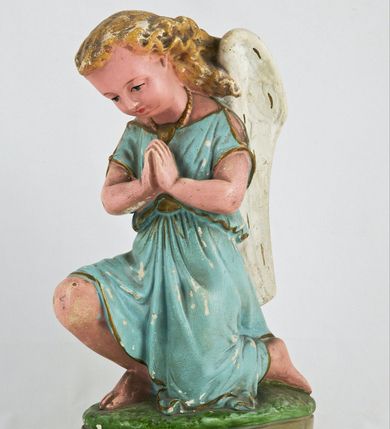 Zdjęcie nr 1: Rzeźba przedstawia anioła klęczącego na lewym kolanie na cokole imitującym rosnącą trawę. Postać ma ręce złożone do modlitwy i opuszczoną głowę z szeroką twarzą o jasnych oczach, małym nosie i czerwonych, niewielkich ustach; jasne włosy opadają jej na plecy. Jest ubrana w niebieską suknię ze złotą lamówką i kołnierzykiem w kropki; skrzydła białe, złożone. 