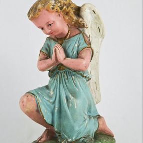 Zdjęcie nr 1: Rzeźba przedstawia anioła klęczącego na lewym kolanie na cokole imitującym rosnącą trawę. Postać ma ręce złożone do modlitwy i opuszczoną głowę z szeroką twarzą o jasnych oczach, małym nosie i czerwonych, niewielkich ustach; jasne włosy opadają jej na plecy. Jest ubrana w niebieską suknię ze złotą lamówką i kołnierzykiem w kropki; skrzydła białe, złożone. 