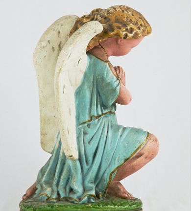 Zdjęcie nr 6: Rzeźba przedstawia anioła klęczącego na lewym kolanie na cokole imitującym rosnącą trawę. Postać ma ręce złożone do modlitwy i opuszczoną głowę z szeroką twarzą o jasnych oczach, małym nosie i czerwonych, niewielkich ustach; jasne włosy opadają jej na plecy. Jest ubrana w niebieską suknię ze złotą lamówką i kołnierzykiem w kropki; skrzydła białe, złożone. 