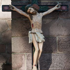 Zdjęcie nr 1: Pełnoplastyczna, polichromowana figura Chrystusa na krzyżu. Sylwetka zmarłego jest smukła, o naturalnej karnacji,  z wyraźnie zaznaczoną muskulaturą i układem kostnym. Ramiona rozpostarte, uniesione na wysokość głowy, nogi są lekko ugięte w kolanach, przechylone na prawą stronę. Głowa opada nisko na prawe ramię, twarz pociągła, oczy zamknięte, nos o prostym profilu, usta lekko otwarte. Włosy ciemne, spływają na plecy i prawe ramię, twarz okala krótka broda. Na głowie ciemnozielona korona cierniowa, biodra otoczone białym, krótkim perizonium podtrzymywanym jasnobrązowym sznurem, opadające pionowymi fałdami z przodu i na prawy bok. Na ciele Chrystusa widoczne są ślady męki: przebity bok, dłonie oraz stopy. Nad głową titulus z napisem „INRI”. Krzyż obwiedziony czarnym profilem, z brązowym wypełnieniem, dekorowany zielonymi liśćmi za głową Chrystusa, wicią wokół plakiety, rozetami na końcach ramion oraz kwitnącą wicią u podstawy.  