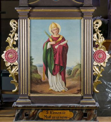 Zdjęcie nr 1: Feretron drewniany w prostej złoconej i malowanej ramie. Pole obrazowe ujęte dwoma kanelowanymi pilastrami. Zwieńczenie w formie łuku kotarowego z krzyżem na osi; w nim medalion z popiersiem Marii, poniżej dwa skrzyżowane młotki. Po bokach rzeźbione uszy z wici roślinnej i rozwiniętych kwiatów. Na awersie całopostaciowe przedstawienie św. Barbary na tle pejzażu, z wieżą po lewej stronie. Święta ukazana została w żółtej sukni, okryta niebieskim, podbitym na różowo, spiętym pod szyją płaszczem, z koroną na głowie. Lewą ręką przytrzymuje oparty o ziemię miecz, a prawą księgę oraz kielich z hostią. Na podstawie feretronu napis „Ś(więta) Barbaro / Módl się za nami”. 
Na rewersie całopostaciowe przedstawienie św. Klemensa na tle pejzażu. Święty został ukazany albie czerwonym ornacie podbitym zielenią, z tiarą na głowie i paliuszem na piersi. W prawej ręce trzyma liść palmowy, a w lewej zwój z napisem „AD CORINTHIOS”, wokół głowy ma złoty nimb. Na podstawie napis „Ś(więty) Klemensie / Módl się za nami”.