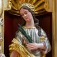 Zdjęcie nr 1: Figura pełna na niskim cokole przedstawiająca nieokreśloną świętą męczennicę. Święta stoi frontalnie z głową przechyloną na prawo, spoglądając delikatnie w dół. Ma podłużną twarz, małe oczy, wąski nos, niewielkie usta i długie, ciemne, spływające falami na ramiona włosy. Prawą rękę wyciąga przed siebie, w lewej trzyma palmę męczeńską. Ubrana jest w srebrną suknię wykończoną przy dekolcie złotą taśmą oraz złoty płaszcz, który owija się wokół jej lewego biodra i przechodzi przez prawą rękę.