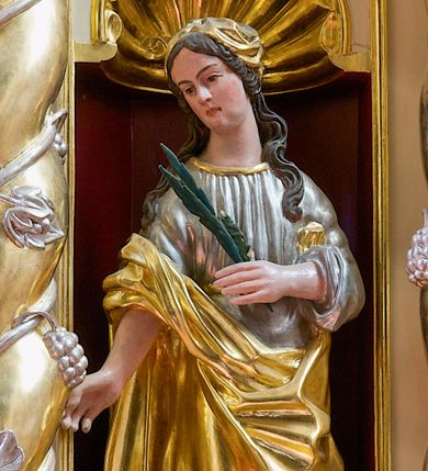 Zdjęcie nr 1: Figura pełna na niskim cokole przedstawiająca nieokreśloną świętą męczennicę. Święta stoi frontalnie z głową przechyloną na prawo, spoglądając delikatnie w dół. Ma podłużną twarz, małe oczy, wąski nos, niewielkie usta i długie, ciemne, spływające falami na ramiona włosy. Prawą rękę wyciąga przed siebie, w lewej trzyma palmę męczeńską. Ubrana jest w srebrną suknię wykończoną przy dekolcie złotą taśmą oraz złoty płaszcz, który owija się wokół jej lewego biodra i przechodzi przez prawą rękę.