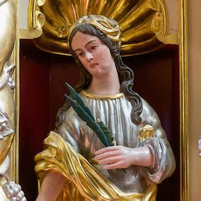 Zdjęcie nr 1: Figura pełna na niskim cokole przedstawiająca nieokreśloną świętą męczennicę. Święta stoi frontalnie z głową przechyloną na prawo, spoglądając delikatnie w dół. Ma podłużną twarz, małe oczy, wąski nos, niewielkie usta i długie, ciemne, spływające falami na ramiona włosy. Prawą rękę wyciąga przed siebie, w lewej trzyma palmę męczeńską. Ubrana jest w srebrną suknię wykończoną przy dekolcie złotą taśmą oraz złoty płaszcz, który owija się wokół jej lewego biodra i przechodzi przez prawą rękę.