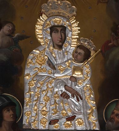 Zdjęcie nr 11: Obraz o kształcie stojącego prostokąta zamkniętego łukiem ostrym. Pośrodku, wśród obłoków przedstawienie Matki Boskiej z Dzieciątkiem na lewym ramieniu, w półpostaci, zwrócona lekko w lewo, z dłońmi skrzyżowanymi w okolicach kolan Chrystusa. Po bokach adorujący dwaj aniołowie Poniżej w narożach kompozycji klęczą po stronie lewej św. Jerzy, po prawej św. Wojciech. Ponad Marią Oko Opatrzności. Matka Boska ubrana jest w czerwoną suknię ze złotą lamówką, na która narzucony ma błękitny płaszcz z kwiatami o żółtych środkach. Głowę wieńczy dekoracyjna korona, podobnie jak Jezusa. Chrystus ukazany jest en trois quarts, w swobodnej pozycji, lewą ręką podtrzymuje  księgę włożoną pod pachę, prawą unosi w geście błogosławieństwa. Ubrany jest w długą sukienkę z długimi rękawami, przewiązaną paskiem, odsłaniającą stopy.  Św. Jerzy, w zbroi i czerwonym płaszczu narzuconym na ramiona, prawą rękę przyłożył do klatki piersiowej, lewą unosi w okolicy tułowia. Na głowie ma hełm z pióropuszem, spod którego widać długie, kręcone włosy opadające na kark. Przy lewym jego boku widoczna jest głowa zabitego smoka. Św. Wojciech, w stroju pontyfikalnym, z paliuszem na ramionach, prawą rękę położył na klatce piersiowej, lewą podtrzymuje połę kapy liturgicznej. Wokół głów świętych koliste nimby. 