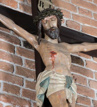Zdjęcie nr 2: Rzeźba pełnoplastyczna, polichromowana. Chrystus w niezbyt głębokim zwisie o sztywnych, mocno napiętych, rozpostartych ramionach i delikatnie zwiniętych palcach. Nogi wyprostowane, skierowane do wewnątrz, dosyć duże stopy nałożone prawa na lewą i przybite jednym gwoździem. Na biodrach krótkie perizonium, z przewagą fałd pionowych, opada na prawym boku kaskadą, z kolei drugi koniec wywinięty na perizonium i spływa na lewe udo. Głowa lekko pochylona w kierunku prawego ramienia, oczy zamknięte, brwi opadające w kierunku skroni, nos prosty, długi, twarz pociągła o wyraźnych kościach policzkowych, usta wąskie, lekko uchylone, okolone przez schematycznie opracowaną w symetryczne rowki brodę. Na głowie korona cierniowa w formie dwóch wałków z licznymi kolcami, spod której opada na prawe ramię cienkie pasmo schematycznie rytych włosów. Z miejsc po przebiciu gwoździami, z rany zadanej w boku oraz spod korony cierniowej spływa krew. Karnacja Chrystusa dosyć jasna, z odcieniem szarości. Krzyż, zarost, włosy, korona cierniowa w kolorze ciemnobrązowym, perizonium białe. W górnej części pionowej belki krzyża zawieszona w pionie tabliczka, dosyć długa, o zwiniętych do wewnątrz końcach, z trójkątnymi wycięciami na środku brzegów, z napisem „I.N.R.I”.