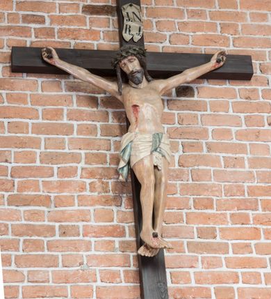 Zdjęcie nr 5: Rzeźba pełnoplastyczna, polichromowana. Chrystus w niezbyt głębokim zwisie o sztywnych, mocno napiętych, rozpostartych ramionach i delikatnie zwiniętych palcach. Nogi wyprostowane, skierowane do wewnątrz, dosyć duże stopy nałożone prawa na lewą i przybite jednym gwoździem. Na biodrach krótkie perizonium, z przewagą fałd pionowych, opada na prawym boku kaskadą, z kolei drugi koniec wywinięty na perizonium i spływa na lewe udo. Głowa lekko pochylona w kierunku prawego ramienia, oczy zamknięte, brwi opadające w kierunku skroni, nos prosty, długi, twarz pociągła o wyraźnych kościach policzkowych, usta wąskie, lekko uchylone, okolone przez schematycznie opracowaną w symetryczne rowki brodę. Na głowie korona cierniowa w formie dwóch wałków z licznymi kolcami, spod której opada na prawe ramię cienkie pasmo schematycznie rytych włosów. Z miejsc po przebiciu gwoździami, z rany zadanej w boku oraz spod korony cierniowej spływa krew. Karnacja Chrystusa dosyć jasna, z odcieniem szarości. Krzyż, zarost, włosy, korona cierniowa w kolorze ciemnobrązowym, perizonium białe. W górnej części pionowej belki krzyża zawieszona w pionie tabliczka, dosyć długa, o zwiniętych do wewnątrz końcach, z trójkątnymi wycięciami na środku brzegów, z napisem „I.N.R.I”.