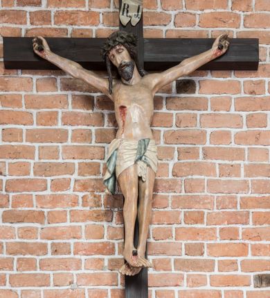 Zdjęcie nr 6: Rzeźba pełnoplastyczna, polichromowana. Chrystus w niezbyt głębokim zwisie o sztywnych, mocno napiętych, rozpostartych ramionach i delikatnie zwiniętych palcach. Nogi wyprostowane, skierowane do wewnątrz, dosyć duże stopy nałożone prawa na lewą i przybite jednym gwoździem. Na biodrach krótkie perizonium, z przewagą fałd pionowych, opada na prawym boku kaskadą, z kolei drugi koniec wywinięty na perizonium i spływa na lewe udo. Głowa lekko pochylona w kierunku prawego ramienia, oczy zamknięte, brwi opadające w kierunku skroni, nos prosty, długi, twarz pociągła o wyraźnych kościach policzkowych, usta wąskie, lekko uchylone, okolone przez schematycznie opracowaną w symetryczne rowki brodę. Na głowie korona cierniowa w formie dwóch wałków z licznymi kolcami, spod której opada na prawe ramię cienkie pasmo schematycznie rytych włosów. Z miejsc po przebiciu gwoździami, z rany zadanej w boku oraz spod korony cierniowej spływa krew. Karnacja Chrystusa dosyć jasna, z odcieniem szarości. Krzyż, zarost, włosy, korona cierniowa w kolorze ciemnobrązowym, perizonium białe. W górnej części pionowej belki krzyża zawieszona w pionie tabliczka, dosyć długa, o zwiniętych do wewnątrz końcach, z trójkątnymi wycięciami na środku brzegów, z napisem „I.N.R.I”.
