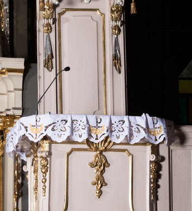 Zdjęcie nr 2: Ambona przyścienna, wisząca, z dostępem w postaci dobudowanych do kosza jednobiegowych schodów z podestem i pełną balustradą, po prawej stronie. Kosz na planie kwadratu o ściętych narożach, na cokole w formie spłaszczonego wałka, z podwieszeniem w postaci ostrosłupa o wklęsłych bokach dekorowanych liśćmi, w narożach ujętego taśmami, zakończonego szyszką. Kosz z pilastrami w narożach, których trzony przechodzą w spływy wolutowe, w partii cokołu przyjmując kształt cęgi, dekorowane są kampanulami i zwisami roślinnymi. Pilastry podtrzymują belkowanie, którego gzyms tworzy parapet kosza. Ścianki zdobione płycinami o kształcie zbliżonym do kwadratu o wklęsłych narożach, zwieńczonymi muszlami z kampanulami. Zaplecek wąski, wydłużony, flankowany lizenami zakończonymi konsolami, dekorowanymi akantem z podwieszonymi pąkami i tkaniną. W polu płycina o kształcie stojącego prostokąta o wklęsłych narożach. Baldachim na planie kwadratu o ściętych narożach, z dołączonymi w ich miejsce impostami z podwieszonymi chwostami. Na frontowej ściance zakończonej dwoma wolutowymi fragmentami gzymsu kartusz z malowanym wizerunkiem niezidentyfikowanego herbu (w czerwonym polu srebrny orzeł z gwiazdą nad głową). Na krawędzi kartusza sygle „K” i „PI”. Kartusz ujęty zwisami kwiatowymi. Na baldachimie cztery kabłąki podtrzymujące wazon, dekorowane kampanulami i ornamentem roślinnym. Na podniebieniu gołębica Ducha Świętego w glorii promienistej. Struktura polichromowana w kolorze białym, profile i ornamentyka złocone, detal srebrzony. 


