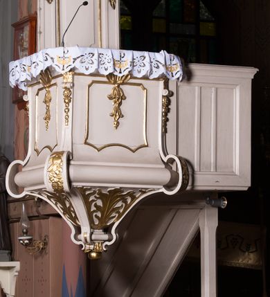 Zdjęcie nr 4: Ambona przyścienna, wisząca, z dostępem w postaci dobudowanych do kosza jednobiegowych schodów z podestem i pełną balustradą, po prawej stronie. Kosz na planie kwadratu o ściętych narożach, na cokole w formie spłaszczonego wałka, z podwieszeniem w postaci ostrosłupa o wklęsłych bokach dekorowanych liśćmi, w narożach ujętego taśmami, zakończonego szyszką. Kosz z pilastrami w narożach, których trzony przechodzą w spływy wolutowe, w partii cokołu przyjmując kształt cęgi, dekorowane są kampanulami i zwisami roślinnymi. Pilastry podtrzymują belkowanie, którego gzyms tworzy parapet kosza. Ścianki zdobione płycinami o kształcie zbliżonym do kwadratu o wklęsłych narożach, zwieńczonymi muszlami z kampanulami. Zaplecek wąski, wydłużony, flankowany lizenami zakończonymi konsolami, dekorowanymi akantem z podwieszonymi pąkami i tkaniną. W polu płycina o kształcie stojącego prostokąta o wklęsłych narożach. Baldachim na planie kwadratu o ściętych narożach, z dołączonymi w ich miejsce impostami z podwieszonymi chwostami. Na frontowej ściance zakończonej dwoma wolutowymi fragmentami gzymsu kartusz z malowanym wizerunkiem niezidentyfikowanego herbu (w czerwonym polu srebrny orzeł z gwiazdą nad głową). Na krawędzi kartusza sygle „K” i „PI”. Kartusz ujęty zwisami kwiatowymi. Na baldachimie cztery kabłąki podtrzymujące wazon, dekorowane kampanulami i ornamentem roślinnym. Na podniebieniu gołębica Ducha Świętego w glorii promienistej. Struktura polichromowana w kolorze białym, profile i ornamentyka złocone, detal srebrzony. 


