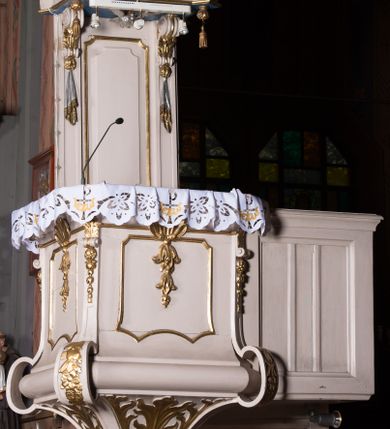 Zdjęcie nr 1: Ambona przyścienna, wisząca, z dostępem w postaci dobudowanych do kosza jednobiegowych schodów z podestem i pełną balustradą, po prawej stronie. Kosz na planie kwadratu o ściętych narożach, na cokole w formie spłaszczonego wałka, z podwieszeniem w postaci ostrosłupa o wklęsłych bokach dekorowanych liśćmi, w narożach ujętego taśmami, zakończonego szyszką. Kosz z pilastrami w narożach, których trzony przechodzą w spływy wolutowe, w partii cokołu przyjmując kształt cęgi, dekorowane są kampanulami i zwisami roślinnymi. Pilastry podtrzymują belkowanie, którego gzyms tworzy parapet kosza. Ścianki zdobione płycinami o kształcie zbliżonym do kwadratu o wklęsłych narożach, zwieńczonymi muszlami z kampanulami. Zaplecek wąski, wydłużony, flankowany lizenami zakończonymi konsolami, dekorowanymi akantem z podwieszonymi pąkami i tkaniną. W polu płycina o kształcie stojącego prostokąta o wklęsłych narożach. Baldachim na planie kwadratu o ściętych narożach, z dołączonymi w ich miejsce impostami z podwieszonymi chwostami. Na frontowej ściance zakończonej dwoma wolutowymi fragmentami gzymsu kartusz z malowanym wizerunkiem niezidentyfikowanego herbu (w czerwonym polu srebrny orzeł z gwiazdą nad głową). Na krawędzi kartusza sygle „K” i „PI”. Kartusz ujęty zwisami kwiatowymi. Na baldachimie cztery kabłąki podtrzymujące wazon, dekorowane kampanulami i ornamentem roślinnym. Na podniebieniu gołębica Ducha Świętego w glorii promienistej. Struktura polichromowana w kolorze białym, profile i ornamentyka złocone, detal srebrzony. 


