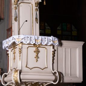 Zdjęcie nr 1: Ambona przyścienna, wisząca, z dostępem w postaci dobudowanych do kosza jednobiegowych schodów z podestem i pełną balustradą, po prawej stronie. Kosz na planie kwadratu o ściętych narożach, na cokole w formie spłaszczonego wałka, z podwieszeniem w postaci ostrosłupa o wklęsłych bokach dekorowanych liśćmi, w narożach ujętego taśmami, zakończonego szyszką. Kosz z pilastrami w narożach, których trzony przechodzą w spływy wolutowe, w partii cokołu przyjmując kształt cęgi, dekorowane są kampanulami i zwisami roślinnymi. Pilastry podtrzymują belkowanie, którego gzyms tworzy parapet kosza. Ścianki zdobione płycinami o kształcie zbliżonym do kwadratu o wklęsłych narożach, zwieńczonymi muszlami z kampanulami. Zaplecek wąski, wydłużony, flankowany lizenami zakończonymi konsolami, dekorowanymi akantem z podwieszonymi pąkami i tkaniną. W polu płycina o kształcie stojącego prostokąta o wklęsłych narożach. Baldachim na planie kwadratu o ściętych narożach, z dołączonymi w ich miejsce impostami z podwieszonymi chwostami. Na frontowej ściance zakończonej dwoma wolutowymi fragmentami gzymsu kartusz z malowanym wizerunkiem niezidentyfikowanego herbu (w czerwonym polu srebrny orzeł z gwiazdą nad głową). Na krawędzi kartusza sygle „K” i „PI”. Kartusz ujęty zwisami kwiatowymi. Na baldachimie cztery kabłąki podtrzymujące wazon, dekorowane kampanulami i ornamentem roślinnym. Na podniebieniu gołębica Ducha Świętego w glorii promienistej. Struktura polichromowana w kolorze białym, profile i ornamentyka złocone, detal srebrzony. 


