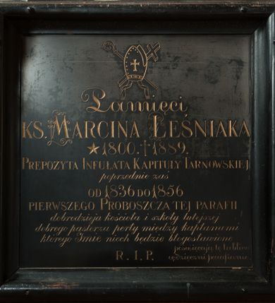 Zdjęcie nr 1: Płyta kamienna w formie kwadratu ujęta drewnianą, profilowaną ramą, polichromowaną na czarno. W polu złocona inskrypcja: „Pamięci / KS(IĘDZA) MARCINA LEŚNIAKA / *1800. +1889. / PREPOZYTA INFUŁATA KAPITUŁY TARNOWSKIEJ / poprzednio zaś / od 1836 do 1856 / PIERWSZEGO PROBOSZCZA TEJ PARAFII / dobrodzieja kościoła i szkoły tutejszej / dobrego pasterza perły między kapłanami / którego Imie niech będzie błogosławione / poświęcają tę tablice / wdzięczni parafianie. / R(equiescat) I(n) P(ace)”. Inicjały imienia i nazwiska zdobione wolutami. Nad inskrypcją ryte przedstawienie infuły z taśmami (fannones), ze skrzyżowanym pastorałem i krzyżem.