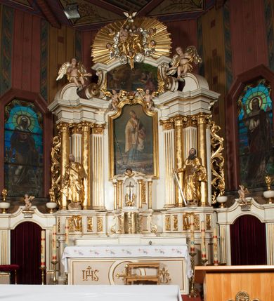 Zdjęcie nr 2: Nastawa architektoniczna, jednokondygnacyjna ze zwieńczeniem, jednoosiowa z bramkami, ustawiona na dwustrefowym cokole, z wyłamanymi pod kątem dwiema parami prostopadłościanów w górnej strefie. Ołtarz kamienny, prostopadłościenny, w drewnianej obudowie, z drewnianą mensą. Antepedium ujęte profilowaną ramą, w centrum medalion z krzyżem o trójlistnym zakończeniu ramion i promieniami na ich skrzyżowaniu, po bokach monogramy maryjny i IHS. Na ołtarzu tabernakulum z tronem wystawienia. Nastawa ujęta dwiema parami kolumn, przedzielonych dwiema parami filarów o kanelowanych trzonach, ustawionych pod kątem. Podpory dźwigają pełne belkowanie, wyłamane pod kątem na ich osi, przerwane nad polem środkowym oraz fragmenty przerwanego, półkolistego przyczółka z siedzącymi na nich puttami. Pomiędzy kolumnami ustawione figury św. Piotra i św. Pawła. W polu środkowym wnęka z obrazem św. Klemensa w formie stojącego prostokąta zakończona łukiem segmentowym z uskokiem, ujęta profilowaną ramą zwieńczoną dwiema zwróconymi ku sobie wolutami, podtrzymującymi koronę otwartą, flankowaną przez dwa putta. Kondygnacja ujęta uszami utworzonymi z suchego akantu i wstęgi. Zwieńczenie o kształcie zbliżonym do leżącego prostokąta, zamkniętego fragmentem segmentowo wyłamanego gzymsu, flankowanego spływami wolutowymi. Całe pole wypełnia obraz Zwiastowania, ujęty liśćmi. Zwieńczenie zakończone płaskorzeźbionym przedstawieniem Trójcy Świętej pośród obłoków i uskrzydlonych główek anielskich, w glorii promienistej. Bramki flankowane pilastrami o kanelowanych trzonach, podtrzymującymi nadproże wyłamane na kształt łuku kotarowego, zwieńczone kluczami z puttami. Na osi pilastrów ustawione po dwa wazony. Przejścia w formie stojącego prostokąta zamkniętego łukiem segmentowym nadwieszonym. Struktura polichromowana w kolorze białym; kolumny, profile i ornamentyka złocone, detal srebrzony. Z tyłu ołtarza ryta inskrypcja: „I.L.O.Z / 9/5 1845”.