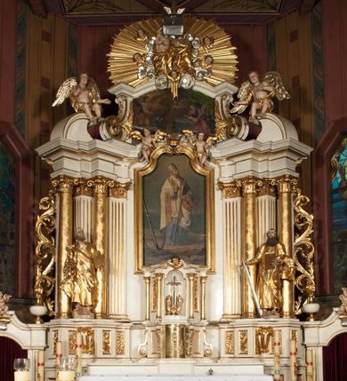Zdjęcie nr 1: Nastawa architektoniczna, jednokondygnacyjna ze zwieńczeniem, jednoosiowa z bramkami, ustawiona na dwustrefowym cokole, z wyłamanymi pod kątem dwiema parami prostopadłościanów w górnej strefie. Ołtarz kamienny, prostopadłościenny, w drewnianej obudowie, z drewnianą mensą. Antepedium ujęte profilowaną ramą, w centrum medalion z krzyżem o trójlistnym zakończeniu ramion i promieniami na ich skrzyżowaniu, po bokach monogramy maryjny i IHS. Na ołtarzu tabernakulum z tronem wystawienia. Nastawa ujęta dwiema parami kolumn, przedzielonych dwiema parami filarów o kanelowanych trzonach, ustawionych pod kątem. Podpory dźwigają pełne belkowanie, wyłamane pod kątem na ich osi, przerwane nad polem środkowym oraz fragmenty przerwanego, półkolistego przyczółka z siedzącymi na nich puttami. Pomiędzy kolumnami ustawione figury św. Piotra i św. Pawła. W polu środkowym wnęka z obrazem św. Klemensa w formie stojącego prostokąta zakończona łukiem segmentowym z uskokiem, ujęta profilowaną ramą zwieńczoną dwiema zwróconymi ku sobie wolutami, podtrzymującymi koronę otwartą, flankowaną przez dwa putta. Kondygnacja ujęta uszami utworzonymi z suchego akantu i wstęgi. Zwieńczenie o kształcie zbliżonym do leżącego prostokąta, zamkniętego fragmentem segmentowo wyłamanego gzymsu, flankowanego spływami wolutowymi. Całe pole wypełnia obraz Zwiastowania, ujęty liśćmi. Zwieńczenie zakończone płaskorzeźbionym przedstawieniem Trójcy Świętej pośród obłoków i uskrzydlonych główek anielskich, w glorii promienistej. Bramki flankowane pilastrami o kanelowanych trzonach, podtrzymującymi nadproże wyłamane na kształt łuku kotarowego, zwieńczone kluczami z puttami. Na osi pilastrów ustawione po dwa wazony. Przejścia w formie stojącego prostokąta zamkniętego łukiem segmentowym nadwieszonym. Struktura polichromowana w kolorze białym; kolumny, profile i ornamentyka złocone, detal srebrzony. Z tyłu ołtarza ryta inskrypcja: „I.L.O.Z / 9/5 1845”.