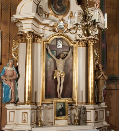 Zdjęcie nr 2: Nastawa architektoniczna, jednoosiowa, jednokondygnacyjna ze zwieńczeniem, ustawiona na dwustrefowym cokole, w górnej części z wyłamanymi pod kątem dwoma prostopadłościanami. Ołtarz drewniany, prostopadłościenny, skrzyniowy, z drewnianą mensą. Antepedium ujęte profilowaną ramą, w polu dwie, duże, płaskorzeźbione rozety na pomarańczowym tle. Nastawa ujęta parą kolumn ustawionych na tle pilastrów, dźwigających pełne belkowanie, wyłamane pod kątem na ich osi, częściowo przerwane nad polem środkowym, oraz fragmenty przerwanego półkolistego przyczółka z puttami. Pole centralne w całości wypełnione wnęką w formie stojącego prostokąta zamkniętego łukiem segmentowym nadwieszonym, ujętym profilowaną ramą, z wpisanym krucyfiksem na tle malowanych obłoków. Nad nim akantowo-wstęgowy kartusz, z napisem „OJCZE! / odpuść im / bo niewiedzą / co czynią”. Kondygnację flankują figury Matki Boskiej i św. Jana ustawione na wysuniętych krawędziach cokołu oraz uszy utworzone z suchego akantu i wstęgi. Zwieńczenie w formie zbliżonej do trapezu zamkniętego fragmentem gzymsu o kształcie łuku segmentowego z uskokiem, flankowanego spływami wolutowymi. W polu obraz Piety w formie stojącego owalu, ujęty profilowaną ramą, u góry dekorowany zwisami kwiatowymi, u dołu liśćmi. Całość zakończona figurą Anioła Stróża. Struktura polichromowana w kolorze białym; kolumny, profile i ornamentyka złocone, detal srebrzony. 
