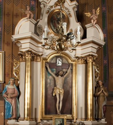 Zdjęcie nr 1: Nastawa architektoniczna, jednoosiowa, jednokondygnacyjna ze zwieńczeniem, ustawiona na dwustrefowym cokole, w górnej części z wyłamanymi pod kątem dwoma prostopadłościanami. Ołtarz drewniany, prostopadłościenny, skrzyniowy, z drewnianą mensą. Antepedium ujęte profilowaną ramą, w polu dwie, duże, płaskorzeźbione rozety na pomarańczowym tle. Nastawa ujęta parą kolumn ustawionych na tle pilastrów, dźwigających pełne belkowanie, wyłamane pod kątem na ich osi, częściowo przerwane nad polem środkowym, oraz fragmenty przerwanego półkolistego przyczółka z puttami. Pole centralne w całości wypełnione wnęką w formie stojącego prostokąta zamkniętego łukiem segmentowym nadwieszonym, ujętym profilowaną ramą, z wpisanym krucyfiksem na tle malowanych obłoków. Nad nim akantowo-wstęgowy kartusz, z napisem „OJCZE! / odpuść im / bo niewiedzą / co czynią”. Kondygnację flankują figury Matki Boskiej i św. Jana ustawione na wysuniętych krawędziach cokołu oraz uszy utworzone z suchego akantu i wstęgi. Zwieńczenie w formie zbliżonej do trapezu zamkniętego fragmentem gzymsu o kształcie łuku segmentowego z uskokiem, flankowanego spływami wolutowymi. W polu obraz Piety w formie stojącego owalu, ujęty profilowaną ramą, u góry dekorowany zwisami kwiatowymi, u dołu liśćmi. Całość zakończona figurą Anioła Stróża. Struktura polichromowana w kolorze białym; kolumny, profile i ornamentyka złocone, detal srebrzony. 