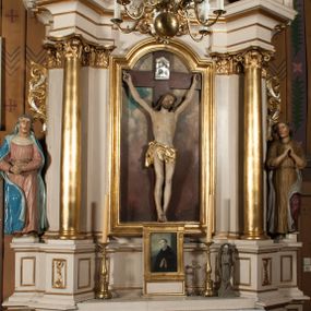 Zdjęcie nr 1: Nastawa architektoniczna, jednoosiowa, jednokondygnacyjna ze zwieńczeniem, ustawiona na dwustrefowym cokole, w górnej części z wyłamanymi pod kątem dwoma prostopadłościanami. Ołtarz drewniany, prostopadłościenny, skrzyniowy, z drewnianą mensą. Antepedium ujęte profilowaną ramą, w polu dwie, duże, płaskorzeźbione rozety na pomarańczowym tle. Nastawa ujęta parą kolumn ustawionych na tle pilastrów, dźwigających pełne belkowanie, wyłamane pod kątem na ich osi, częściowo przerwane nad polem środkowym, oraz fragmenty przerwanego półkolistego przyczółka z puttami. Pole centralne w całości wypełnione wnęką w formie stojącego prostokąta zamkniętego łukiem segmentowym nadwieszonym, ujętym profilowaną ramą, z wpisanym krucyfiksem na tle malowanych obłoków. Nad nim akantowo-wstęgowy kartusz, z napisem „OJCZE! / odpuść im / bo niewiedzą / co czynią”. Kondygnację flankują figury Matki Boskiej i św. Jana ustawione na wysuniętych krawędziach cokołu oraz uszy utworzone z suchego akantu i wstęgi. Zwieńczenie w formie zbliżonej do trapezu zamkniętego fragmentem gzymsu o kształcie łuku segmentowego z uskokiem, flankowanego spływami wolutowymi. W polu obraz Piety w formie stojącego owalu, ujęty profilowaną ramą, u góry dekorowany zwisami kwiatowymi, u dołu liśćmi. Całość zakończona figurą Anioła Stróża. Struktura polichromowana w kolorze białym; kolumny, profile i ornamentyka złocone, detal srebrzony. 