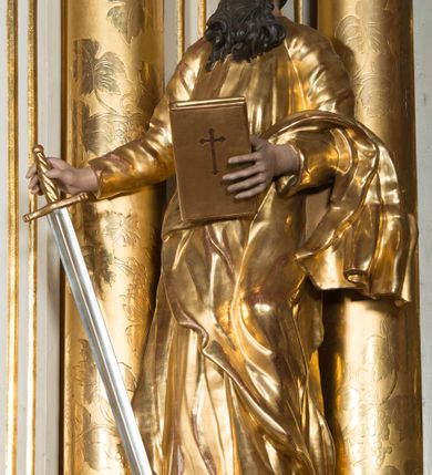 Zdjęcie nr 5: Rzeźba wydrążona z tyłu, ustawiona na nieregularnej podstawie. Święty ukazany frontalnie, w kontrapoście, w prawej, wyciągniętej w bok ręce trzyma miecz zwrócony ostrzem w dół, w lewej, ugiętej, trzyma księgę. Twarz ma szczupłą, kwadratową, o starczych rysach, migdałowatych oczach, garbatym nosie i otwartych ustach z widoczną linią zębów. Twarz ma okoloną długą, silnie kręconą brodą oraz krótkimi, falowanymi włosami. Ubrany jest w złoconą tunikę, przepasaną w talii oraz złocony płaszcz zarzucony na lewe przedramię i bok. Polichromia ciała naturalistyczna, włosy i broda ciemnobrązowe. 

