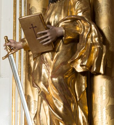 Zdjęcie nr 6: Rzeźba wydrążona z tyłu, ustawiona na nieregularnej podstawie. Święty ukazany frontalnie, w kontrapoście, w prawej, wyciągniętej w bok ręce trzyma miecz zwrócony ostrzem w dół, w lewej, ugiętej, trzyma księgę. Twarz ma szczupłą, kwadratową, o starczych rysach, migdałowatych oczach, garbatym nosie i otwartych ustach z widoczną linią zębów. Twarz ma okoloną długą, silnie kręconą brodą oraz krótkimi, falowanymi włosami. Ubrany jest w złoconą tunikę, przepasaną w talii oraz złocony płaszcz zarzucony na lewe przedramię i bok. Polichromia ciała naturalistyczna, włosy i broda ciemnobrązowe. 

