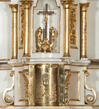 Zdjęcie nr 1: Tabernakulum o kształcie prostopadłościennej skrzynki obitej blachą o wypukłym licu. Drzwiczki w formie stojącego prostokąta z przedstawieniem kielicha z hostią w glorii promienistej oraz winnej latorośli z kłosami zbóż. Tabernakulum w drewnianej obudowie, z dostawionymi po bokach prostopadłościanami, o wklęsłych licach, z parą pilastrów o konsolach z kampanulami w miejscu trzonów, ujęte spływami wolutowymi. Tron wystawienia w formie sześcianu, o ściętych narożach, ujęty czterema kolumienkami podtrzymującymi imposty oraz gzyms wyłamujący się pośrodku na kształt łuku odcinkowego. W polu środkowym obrotowa nisza o wykroju stojącego prostokąta zamkniętego łukiem półkolistym nadwieszonym. Na osiach bocznych płyciny z ornamentem roślinnym. Całość zwieńczona uskrzydloną główką anielską. Struktura polichromowana w kolorze białym, kolumienki, profile i ornamentyka złocone. 