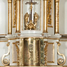 Zdjęcie nr 1: Tabernakulum o kształcie prostopadłościennej skrzynki obitej blachą o wypukłym licu. Drzwiczki w formie stojącego prostokąta z przedstawieniem kielicha z hostią w glorii promienistej oraz winnej latorośli z kłosami zbóż. Tabernakulum w drewnianej obudowie, z dostawionymi po bokach prostopadłościanami, o wklęsłych licach, z parą pilastrów o konsolach z kampanulami w miejscu trzonów, ujęte spływami wolutowymi. Tron wystawienia w formie sześcianu, o ściętych narożach, ujęty czterema kolumienkami podtrzymującymi imposty oraz gzyms wyłamujący się pośrodku na kształt łuku odcinkowego. W polu środkowym obrotowa nisza o wykroju stojącego prostokąta zamkniętego łukiem półkolistym nadwieszonym. Na osiach bocznych płyciny z ornamentem roślinnym. Całość zwieńczona uskrzydloną główką anielską. Struktura polichromowana w kolorze białym, kolumienki, profile i ornamentyka złocone. 