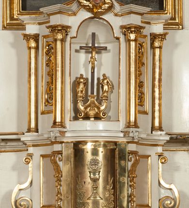 Zdjęcie nr 2: Tabernakulum o kształcie prostopadłościennej skrzynki obitej blachą o wypukłym licu. Drzwiczki w formie stojącego prostokąta z przedstawieniem kielicha z hostią w glorii promienistej oraz winnej latorośli z kłosami zbóż. Tabernakulum w drewnianej obudowie, z dostawionymi po bokach prostopadłościanami, o wklęsłych licach, z parą pilastrów o konsolach z kampanulami w miejscu trzonów, ujęte spływami wolutowymi. Tron wystawienia w formie sześcianu, o ściętych narożach, ujęty czterema kolumienkami podtrzymującymi imposty oraz gzyms wyłamujący się pośrodku na kształt łuku odcinkowego. W polu środkowym obrotowa nisza o wykroju stojącego prostokąta zamkniętego łukiem półkolistym nadwieszonym. Na osiach bocznych płyciny z ornamentem roślinnym. Całość zwieńczona uskrzydloną główką anielską. Struktura polichromowana w kolorze białym, kolumienki, profile i ornamentyka złocone. 