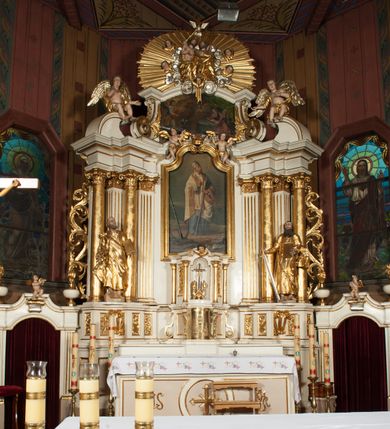 Zdjęcie nr 3: Tabernakulum o kształcie prostopadłościennej skrzynki obitej blachą o wypukłym licu. Drzwiczki w formie stojącego prostokąta z przedstawieniem kielicha z hostią w glorii promienistej oraz winnej latorośli z kłosami zbóż. Tabernakulum w drewnianej obudowie, z dostawionymi po bokach prostopadłościanami, o wklęsłych licach, z parą pilastrów o konsolach z kampanulami w miejscu trzonów, ujęte spływami wolutowymi. Tron wystawienia w formie sześcianu, o ściętych narożach, ujęty czterema kolumienkami podtrzymującymi imposty oraz gzyms wyłamujący się pośrodku na kształt łuku odcinkowego. W polu środkowym obrotowa nisza o wykroju stojącego prostokąta zamkniętego łukiem półkolistym nadwieszonym. Na osiach bocznych płyciny z ornamentem roślinnym. Całość zwieńczona uskrzydloną główką anielską. Struktura polichromowana w kolorze białym, kolumienki, profile i ornamentyka złocone. 