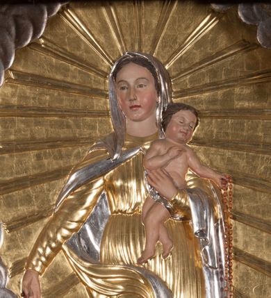 Zdjęcie nr 1: Płaskorzeźba w kształcie stojącego prostokąta o ściętych górnych narożach. W centrum kompozycji na wielobocznym cokole z rytą datą „1872” stoi Maria, ukazana frontalnie, w kontrapoście, lewą ręką podtrzymuje Dzieciątko, prawą wyciąga w bok, w dłoni trzyma różaniec. Twarz ma owalną, o łagodnych rysach, migdałowatych oczach, prostym nosie i pełnych ustach, okoloną brązowymi włosami ukrytymi pod welonem. Ubrana jest w przylegającą do ciała, obficie drapowaną suknię, przepasaną w talii, pod szyją ma zapięty płaszcz opadający na ramiona i prawą nogę, a na głowie welon. Dzieciątko w pozycji siedzącej, o esowato wygiętej sylwetce, zwrócone w lewo, prawą rękę ugina i unosi w geście błogosławieństwa, lewą wyciąga przed siebie, w dłoni trzyma różaniec. Twarz ma kwadratową, o pełnych policzkach, zamkniętych oczach, małym nosie i ustach, okoloną krótkimi, brązowymi włosami. Ciało ma pulchne, w partii bioder przewiązane pieluszką. W lewym dolnym rogu obrazu klęcząca postać św. Dominika, ukazana z prawego profilu, z uniesioną głową i rękami złożonymi w geście modlitwy. Twarz ma szczupłą, z oczami skierowanymi w górę, okoloną krótką brodą i włosami, na głowie ma tonsurę. Ubrany jest w habit ze szkaplerzem oraz płaszcz z kapturem. Przed nim, na ziemi leży pies z pochodnią w pysku. W prawym dolnym rogu klęcząca św. Katarzyna Sieneńska, zwrócona w trzech czwartych do środka kompozycji, z dłońmi złożonymi w geście modlitwy. Twarz ma pociągłą, o zamkniętych oczach, długim nosie i wąskich ustach. Wokół głowy ma podwikę, na niej welon. Ubrana jest w habit ze szkaplerzem, na ramiona ma narzucony płaszcz. Wokół Marii gloria promienista, tło przedstawienia wypełnione obłokami. Polichromia w partiach ciała naturalistyczna, stroje i tło złocone i srebrzone. 