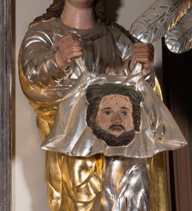 Zdjęcie nr 2: Figura pełnoplastyczna. Anioł ukazany frontalnie, z ugiętą lewą nogą. Obie ręce ma rozłożone na boki, w prawej trzyma włócznię. Twarz ma owalną, o pełnych policzkach, dużych oczach, zadartym nosie i rozchylonych ustach; okoloną falowanymi włosami opadającymi na ramiona. Ubrany jest w srebrzoną suknię spodnią, sięgającą ziemi, odsłaniającą lewą nogę oraz krótszą szatę wierzchnią, złoconą, o szerokich rękawach, przepasaną w talii. Skrzydła ma srebrzone, rozłożone na boki. Polichromia ciała naturalistyczna, włosy brązowe. 

