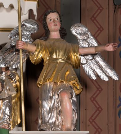 Zdjęcie nr 1: Figura pełnoplastyczna. Anioł ukazany frontalnie, z ugiętą lewą nogą. Obie ręce ma rozłożone na boki, w prawej trzyma włócznię. Twarz ma owalną, o pełnych policzkach, dużych oczach, zadartym nosie i rozchylonych ustach; okoloną falowanymi włosami opadającymi na ramiona. Ubrany jest w srebrzoną suknię spodnią, sięgającą ziemi, odsłaniającą lewą nogę oraz krótszą szatę wierzchnią, złoconą, o szerokich rękawach, przepasaną w talii. Skrzydła ma srebrzone, rozłożone na boki. Polichromia ciała naturalistyczna, włosy brązowe. 


