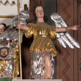 Zdjęcie nr 1: Figura pełnoplastyczna. Anioł ukazany frontalnie, z ugiętą lewą nogą. Obie ręce ma rozłożone na boki, w prawej trzyma włócznię. Twarz ma owalną, o pełnych policzkach, dużych oczach, zadartym nosie i rozchylonych ustach; okoloną falowanymi włosami opadającymi na ramiona. Ubrany jest w srebrzoną suknię spodnią, sięgającą ziemi, odsłaniającą lewą nogę oraz krótszą szatę wierzchnią, złoconą, o szerokich rękawach, przepasaną w talii. Skrzydła ma srebrzone, rozłożone na boki. Polichromia ciała naturalistyczna, włosy brązowe. 

