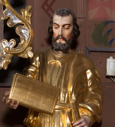 Zdjęcie nr 1: Figura pełnoplastyczna, ustawiona na nieregularnej podstawie. Święty ukazany frontalnie, z lekko pochyloną głową. W prawej, ugiętej ręce trzyma zamkniętą księgę, w lewej pałkę. Twarz ma pociągłą, o dojrzałych rysach, zmarszczonym czole, półprzymkniętych oczach, prostym nosie i pełnych ustach, okoloną krótką, falistą brodą i włosami. Ubrany jest w złoconą tunikę z kołnierzykiem, przepasaną w talii, przylegającą do ciała, silnie drapowaną oraz płaszcz zarzucony na prawe ramię postaci i opadający po bokach. Polichromia ciała naturalistyczna, włosy brązowe. 

