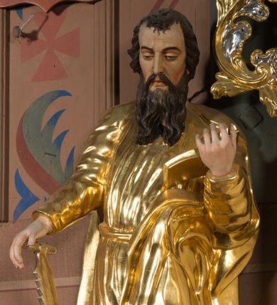 Zdjęcie nr 1: Figura pełnoplastyczna ustawiona na nieregularnej podstawie. Święty ukazany frontalnie, z pochyloną głową, lewą nogą ugiętą, opartą na podstawie. Lewą ręką podtrzymuje otwartą księgę, prawą wyciąga w bok i opiera na pile. Twarz ma pociągłą, o dojrzałych rysach, zmarszczonym czole, półprzymkniętych oczach, prostym nosie i pełnych ustach, okoloną długą, rozdwajającą się brodą i krótkimi, falowanymi włosami. Ubrany jest w złoconą tunikę, przylegającą do ciała, obficie drapowaną, przepasaną w talii oraz płaszcz zarzucony na ramiona i na lewy bok postaci. Polichromia ciała naturalistyczna, włosy brązowe. 
