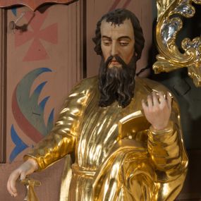 Zdjęcie nr 1: Figura pełnoplastyczna ustawiona na nieregularnej podstawie. Święty ukazany frontalnie, z pochyloną głową, lewą nogą ugiętą, opartą na podstawie. Lewą ręką podtrzymuje otwartą księgę, prawą wyciąga w bok i opiera na pile. Twarz ma pociągłą, o dojrzałych rysach, zmarszczonym czole, półprzymkniętych oczach, prostym nosie i pełnych ustach, okoloną długą, rozdwajającą się brodą i krótkimi, falowanymi włosami. Ubrany jest w złoconą tunikę, przylegającą do ciała, obficie drapowaną, przepasaną w talii oraz płaszcz zarzucony na ramiona i na lewy bok postaci. Polichromia ciała naturalistyczna, włosy brązowe. 
